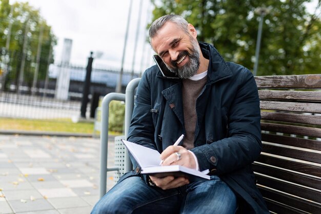 Um homem em um parque da cidade senta-se em um banco, estou falando ao telefone, escreve em um caderno