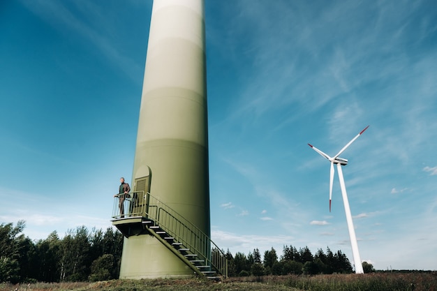 Um homem em um terno com uma camisa de golfe verde fica ao lado de um moinho de vento no contexto do campo e o céu azul. Homem de negócios perto dos moinhos de vento. Conceito moderno do futuro.