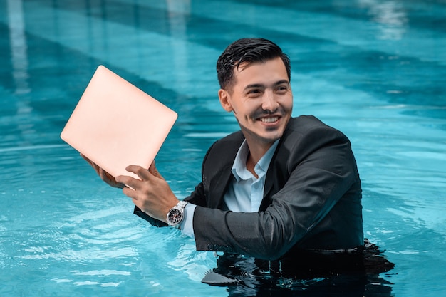 Um homem em um terno de negócios está pronto para afogar seu laptop enquanto está na piscina