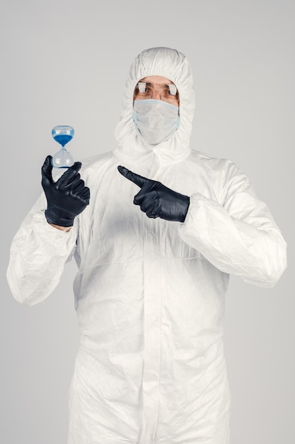 Um homem em um traje de proteção branco uma máscara em um fundo branco com um relógio nas mãos Uma pandemia epidêmica é um novo vírus que se espalha rapidamente um conceito médico