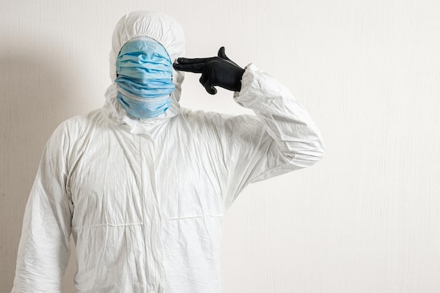 Um homem em um traje de proteção pendurado com máscaras médicas posando contra um fundo de parede mostrando vários gestos com os dedos o cientista aponta uma pistola para o templo