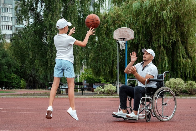 Um homem em uma cadeira de rodas joga basquete com o filho no campo de esportes. Pai com deficiência, infância feliz, conceito de pessoa com deficiência.