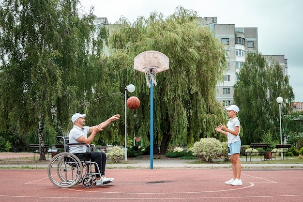 Foto um homem em uma cadeira de rodas joga basquete com o filho no campo de esportes. pai com deficiência, infância feliz, conceito de pessoa com deficiência.