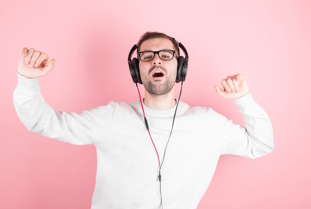 Um homem engraçado com barba e um moletom branco ouve música com fones de ouvido, canta e dança sobre um fundo rosa.