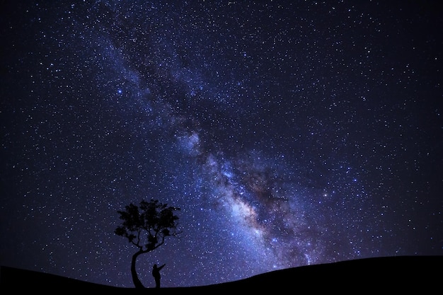Um homem está parado na árvore apontando para uma estrela brilhante com a Via Láctea
