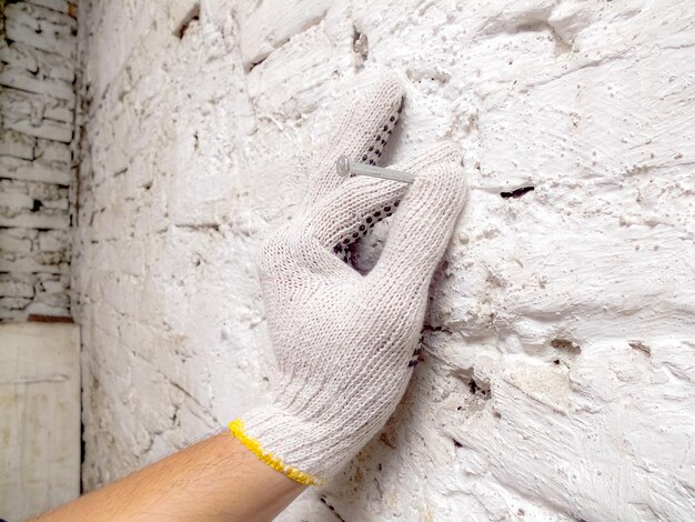 Foto um homem está segurando um prego para ser batido em uma parede de tijolos brancos usando luvas brancas