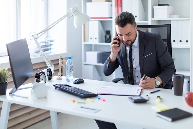 Um homem está sentado à mesa do escritório, falando ao telefone e segurando um marcador na mão.