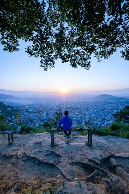 Um homem está sentado em um banco olhando para uma cidade ao pôr do sol.