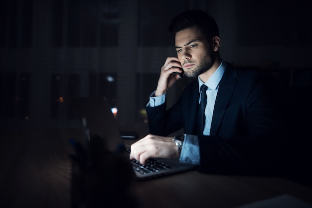 Um homem está sentado em um laptop em um escritório escuro.