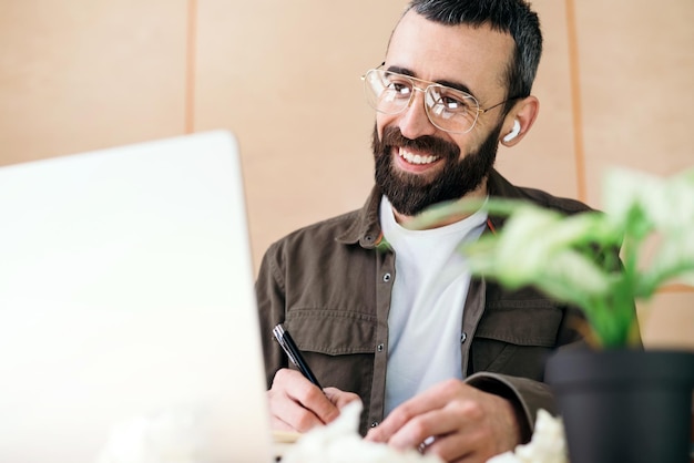 Um homem está sentado em uma mesa com um laptop e uma caneta na mão.