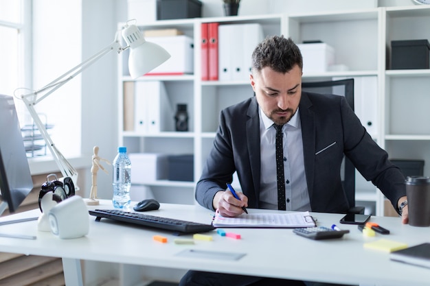 Um homem está sentado no escritório à mesa, segurando um copo de café e uma caneta na mão e trabalhando com documentos