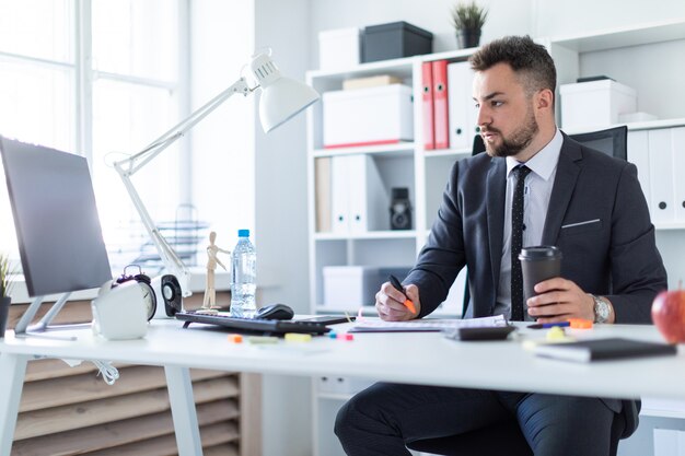 Um homem está sentado no escritório à mesa, segurando um marcador e um copo de café na mão e olhando para o monitor.