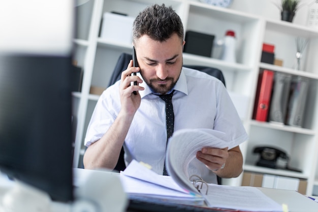 Um homem está sentado no escritório, trabalhando com documentos e falando ao telefone.