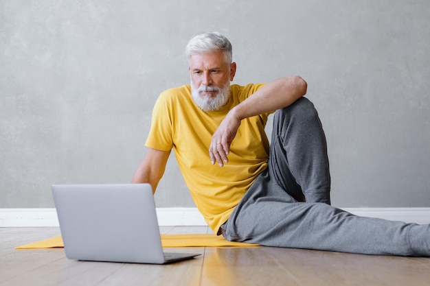 Um homem faz ioga e fitness no ginásio com um treinador on-line de um treinador de esportes masculino sênior de laptop