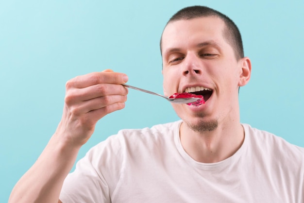 Um homem feliz com a boca aberta comendo uma doce geléia vermelha com uma colher em um fundo azul Desfrutando de deliciosa dieta comida Gelatina Sabor saboroso Fruta Cereja Saudável Berry Sobremesa Branco Vermelho