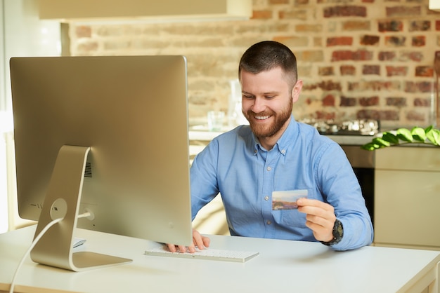 Um homem feliz com barba digita as informações do cartão de crédito para fazer compras on-line em casa