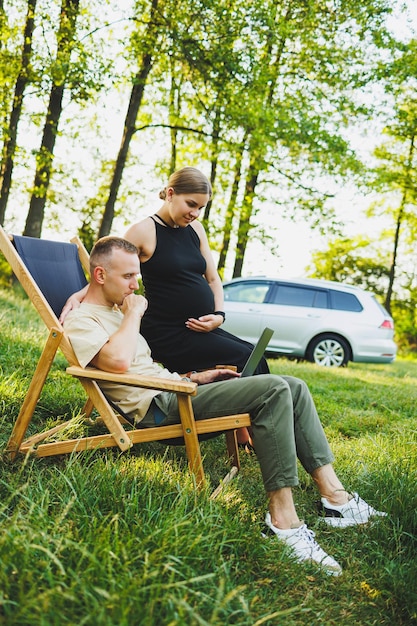 Um homem feliz e sua esposa grávida estão trabalhando em um laptop enquanto estão sentados na natureza Um jovem casal está relaxando na natureza e vendo um filme em um laptop