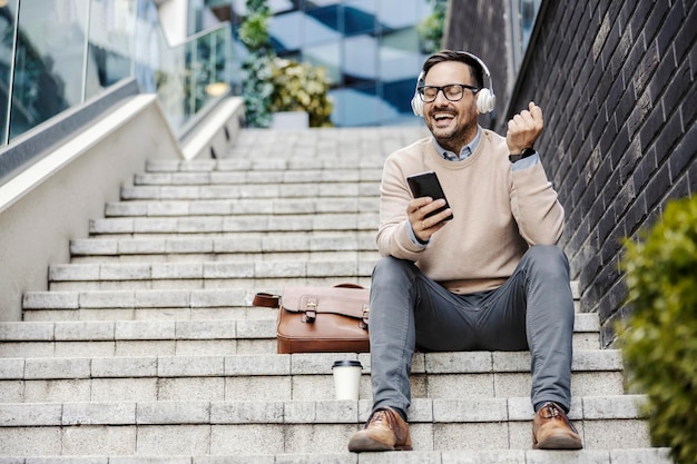 Um homem feliz em casual inteligente está sentado na escada ao ar livre e curtindo música no telefone