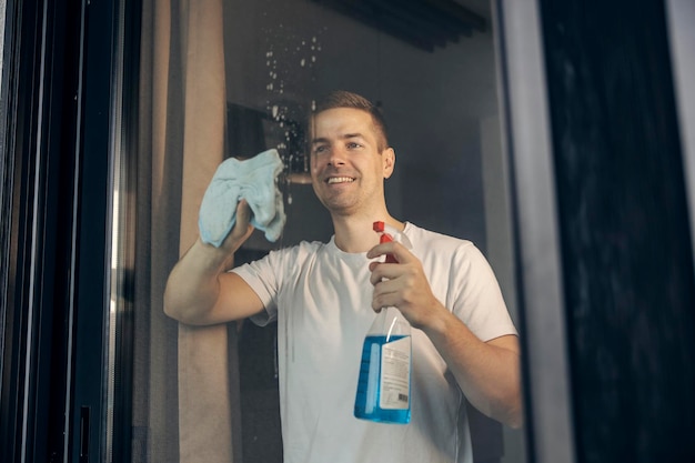 Um homem feliz está borrifando a janela com detergente e esfregando com um pano