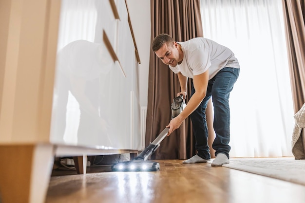 Um homem feliz está fazendo tarefas em casa e aspirando sob os móveis