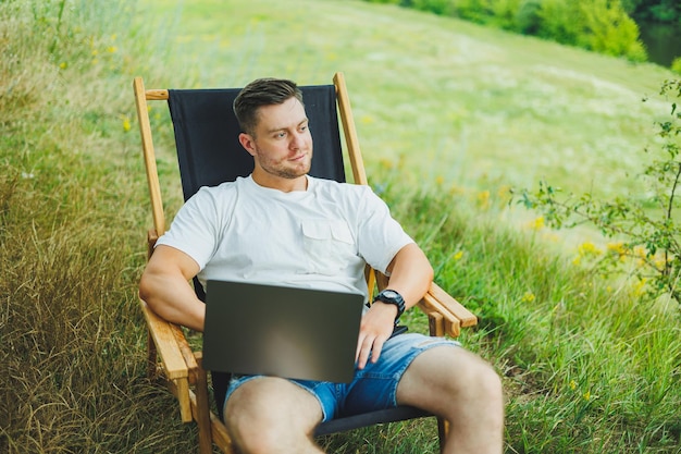 Um homem feliz está relaxando na natureza e trabalhando remotamente em um laptop portátil Trabalho remoto enquanto está de férias Trabalhando em um laptop enquanto está sentado em uma cadeira na natureza