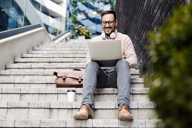 Um homem feliz senta-se na escada do lado de fora e usando laptop