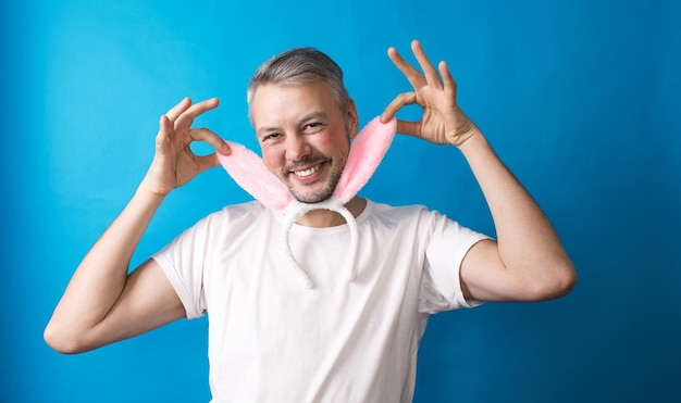 Um homem gay com maquiagem em uma camiseta branca em um fundo azul segura orelhas de coelho em suas mãos olhando para a câmera