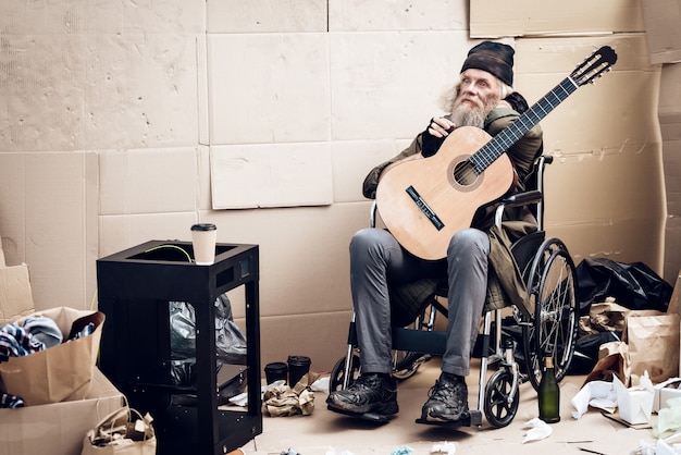 Um homem grisalho com barba senta-se perto de lixo com uma guitarra