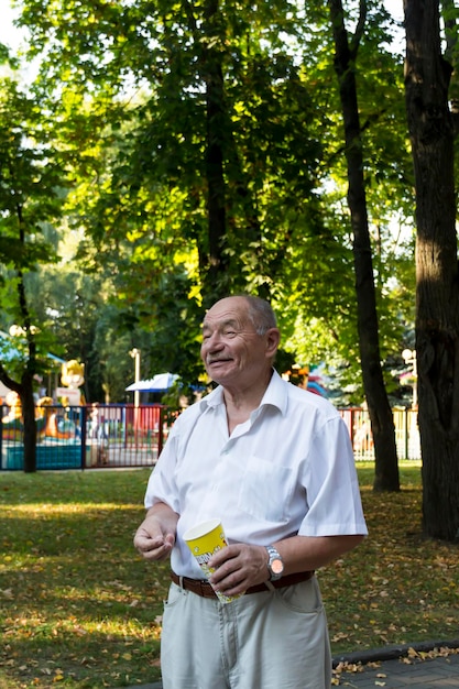 Foto um homem idoso caminha sozinho no parque no verão um aposentado de camisa branca fica sozinho no parque com um copo de pipoca
