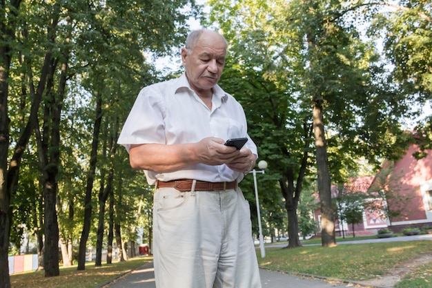 Um homem idoso caminha sozinho no parque no verão Um empresário aposentado moderno em uma camisa branca e calça escreve uma mensagem e estuda informações em um telefone celular