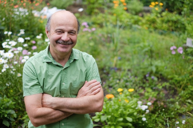 Um homem idoso com bigode e careca em uma camiseta verde está de pé entre flores no jardim de verão braços cruzados e sorrindo