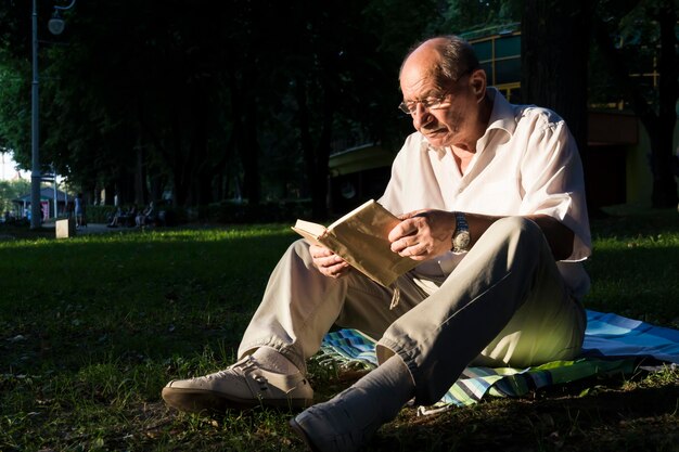 Um homem idoso de camisa branca está sentado em um cobertor no chão de um parque e lendo.