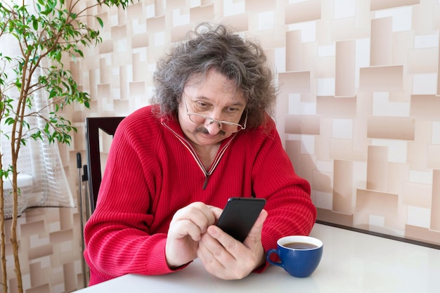 Um homem idoso de suéter vermelho e óculos com um telefone nas mãos está sentado à mesa