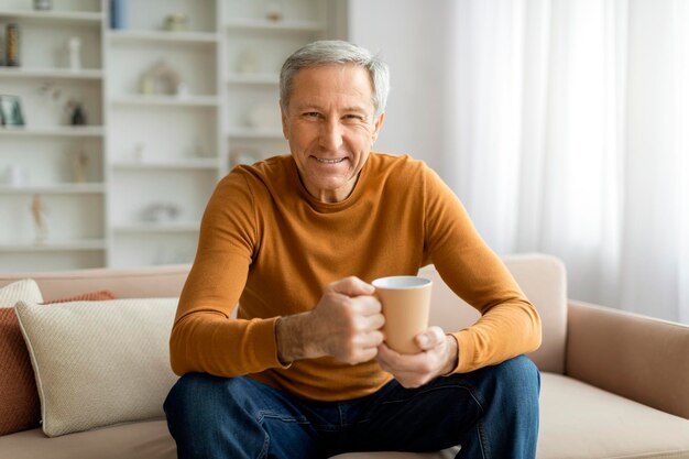 Um homem idoso europeu alegre sentado no sofá a beber café.