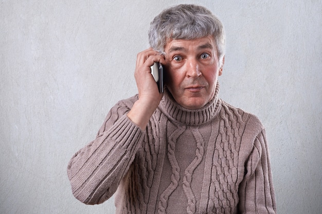 Um homem idoso surpreso na camisola com cabelos grisalhos, olhos escuros e rugas no rosto, comunicando-se por telefone