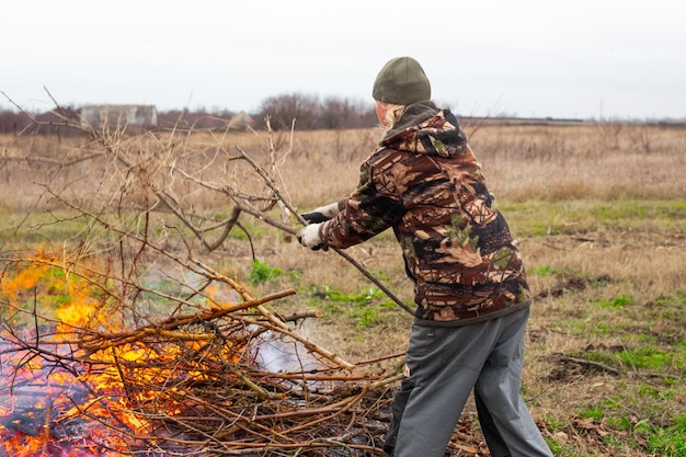 Um homem joga galhos secos em uma fogueira em um campo Queimando lixo e galhos no jardim