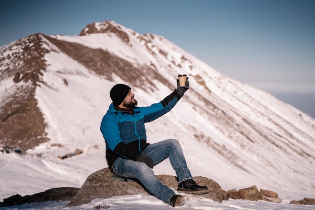 Um homem levanta uma xícara de café no topo de uma montanha de inverno
