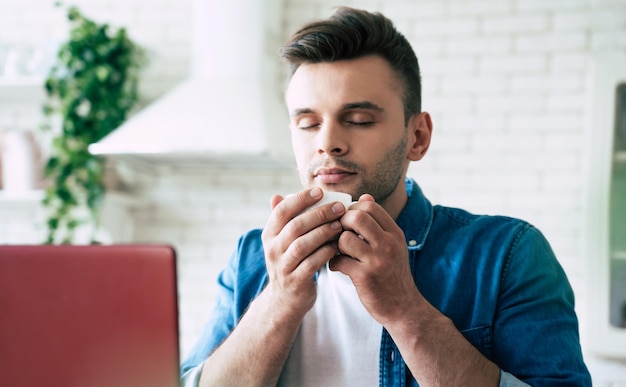 Um homem lindo e sorridente está usando um laptop e bebe café ou chá enquanto está sentado na cozinha de casa