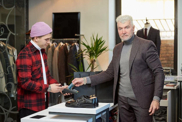 Um homem maduro com cabelos grisalhos e físico esportivo está aplicando um cartão de crédito em um terminal de ponto de venda em uma loja de roupas Um cliente do sexo masculino com barba e um assistente de loja em uma boutique
