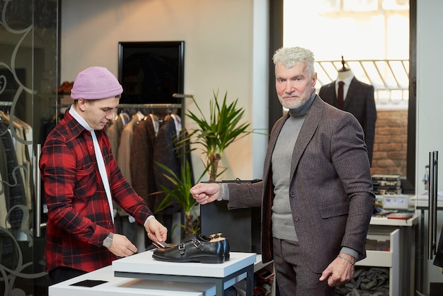 Um homem maduro com cabelos grisalhos e um físico esportivo está posando durante o pagamento de uma compra em uma loja de roupas Um cliente do sexo masculino com barba e um assistente de loja em uma boutique