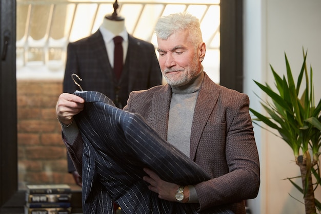 Um homem maduro de cabelos grisalhos e físico esportivo está verificando um terno de lã em uma loja de roupas. Um cliente do sexo masculino com barba em uma boutique.
