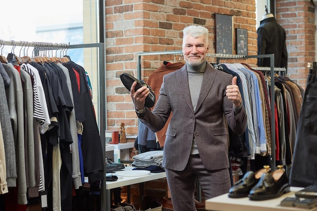 Um homem maduro feliz com cabelos grisalhos e um físico esportivo está posando segurando um sapato preto e mostrando os polegares em uma loja de roupas Um cliente do sexo masculino com barba usa um terno de lã em uma boutique