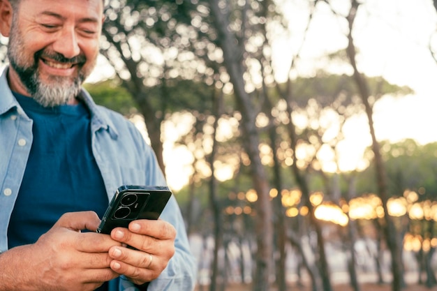 Um homem maduro moderno usando smartphone ao ar livre no parque sorrindo e se divertindo Compartilhamento de vida nas mídias sociais carregando conteúdo Pessoas escrevendo no celular em comunicação de conexão ao ar livre Adulto