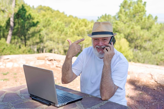 Um homem mais velho gesticulando enquanto trabalhava remotamente em seu laptop em uma mesa ao ar livre e falando em seu celular