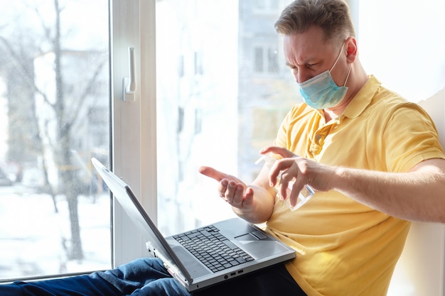 Um homem mascarado desinfeta as mãos antes de trabalhar em um laptop