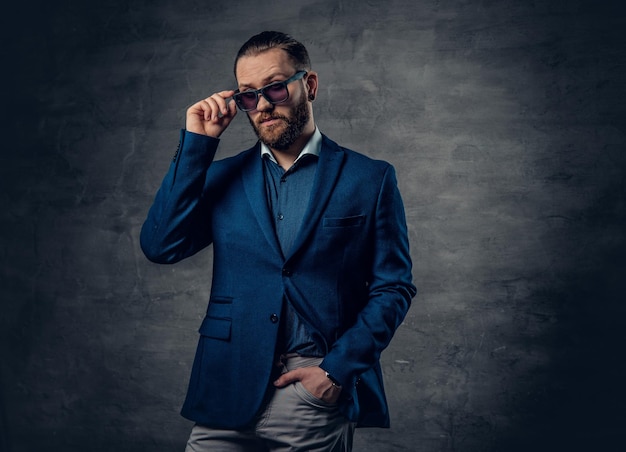 Um homem moderno barbudo elegante vestido de terno e óculos escuros sobre fundo cinza escuro.