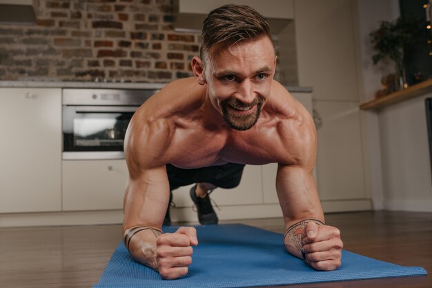 Um homem musculoso com um torso nu está fazendo uma prancha em um tapete de ioga azul em seu apartamento à noite