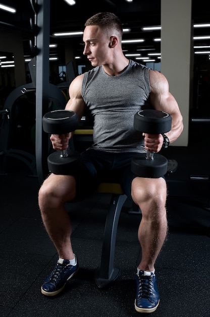 Um homem musculoso está segurando halteres sobre os joelhos em uma academia.