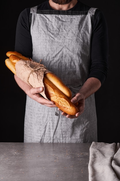 Um homem padeiro com barba em um avental cinza se levanta contra um fundo preto e segura, quebra, corta pão delicioso crocante, pãezinhos, baguete.