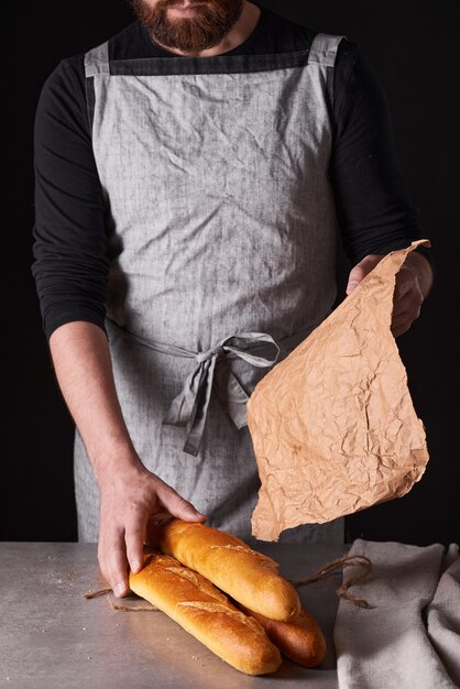 Um homem padeiro com barba em um avental cinza se levanta contra um fundo preto e segura, quebra, corta pão delicioso crocante, pãezinhos, baguete.